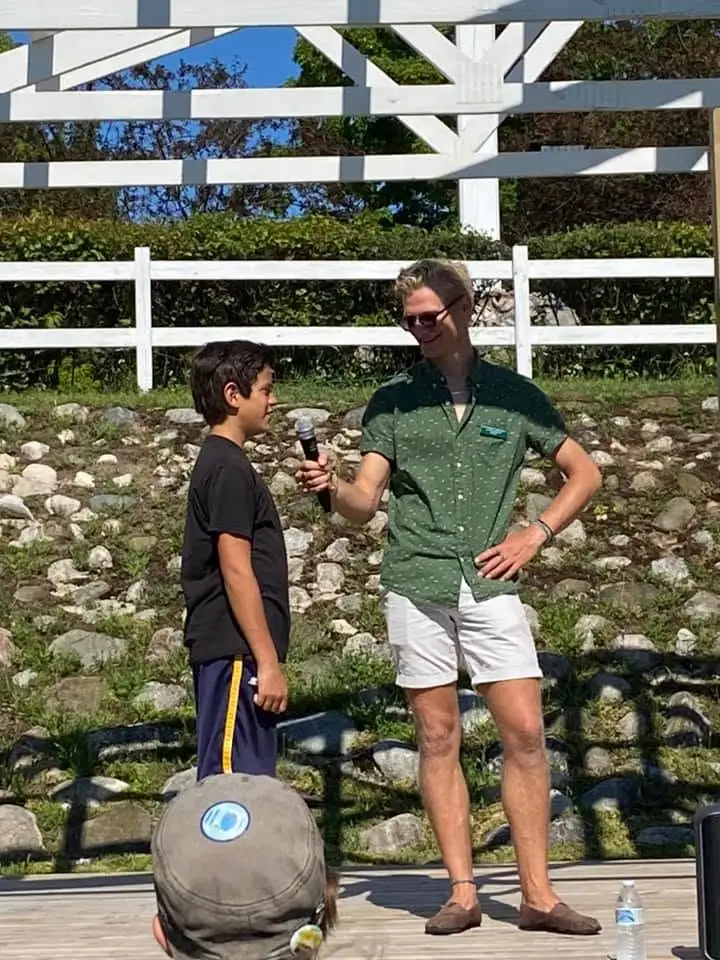 An adult holds a microphone toward a boy outdoors beside a white fence and rocky ground.