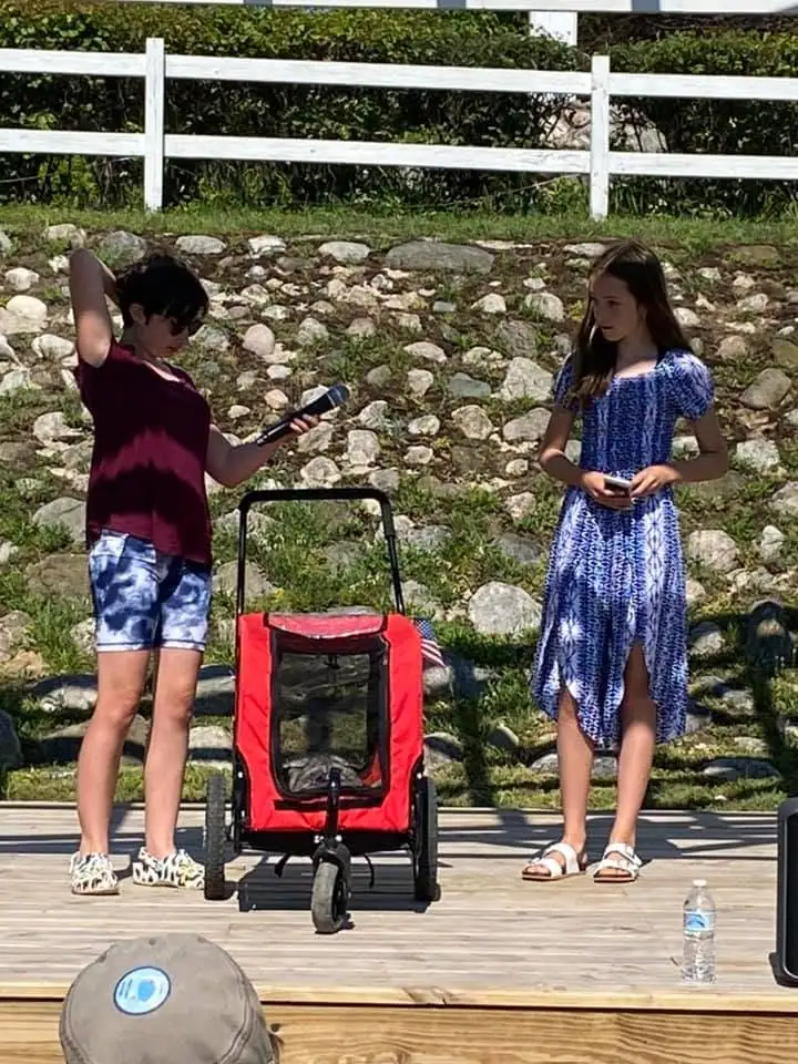 Two young people stand beside a red wagon on a wooden deck, holding a microphone and a phone.