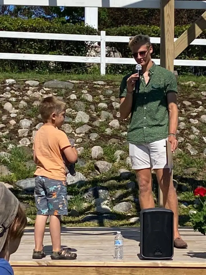 An adult speaks into a microphone beside a child on an outdoor wooden stage.