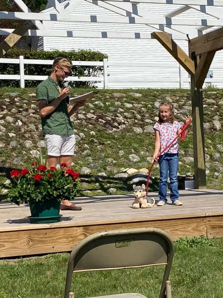 An adult speaks into a microphone beside a child holding a stuffed dog on a leash on an outdoor wooden stage.