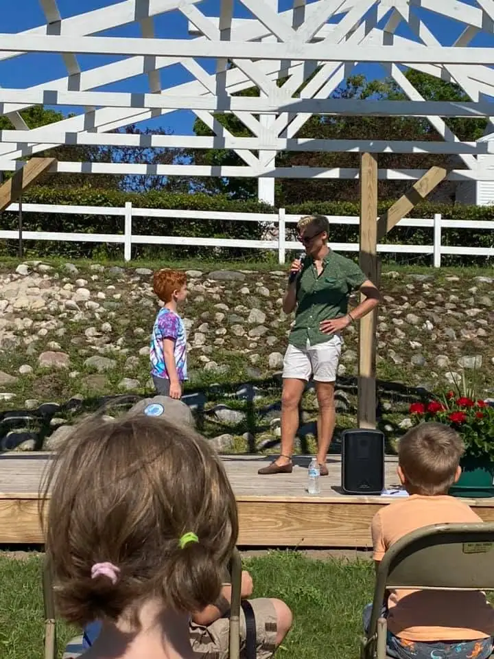 An adult speaks into a microphone to a child on an outdoor wooden stage while seated children watch.