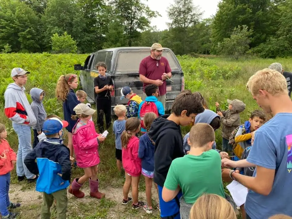 Children and adults gather around a pickup truck during an outdoor activity in a grassy, wooded area.