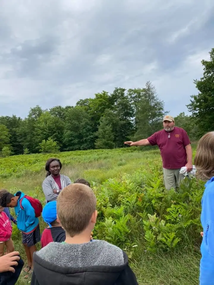 An adult points across dense meadow vegetation while children and another adult watch.