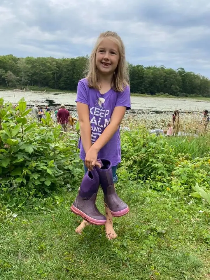 A barefoot girl holds purple rubber boots beside a lily-covered pond.