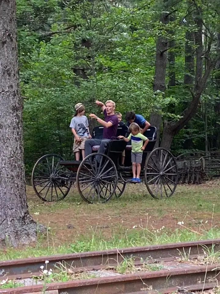 An adult and four children sit and stand on a horse-drawn carriage beside railroad tracks in a wooded setting.