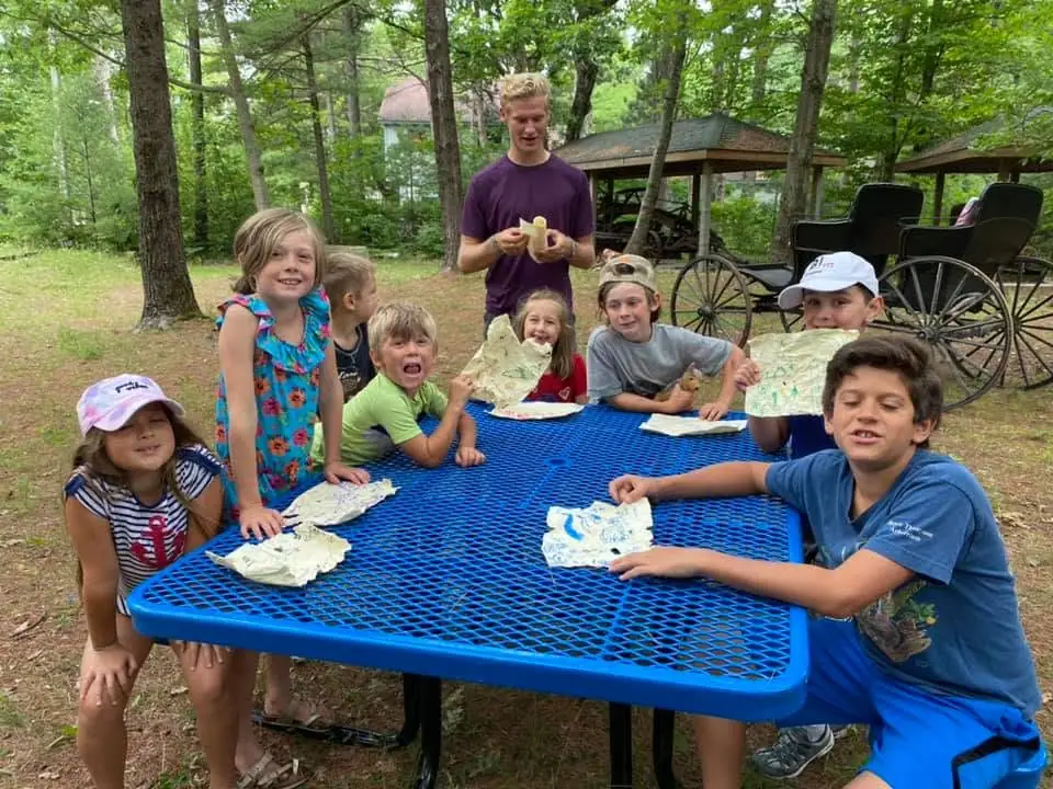 Children and an adult gather around a blue picnic table with maps in a wooded outdoor setting.