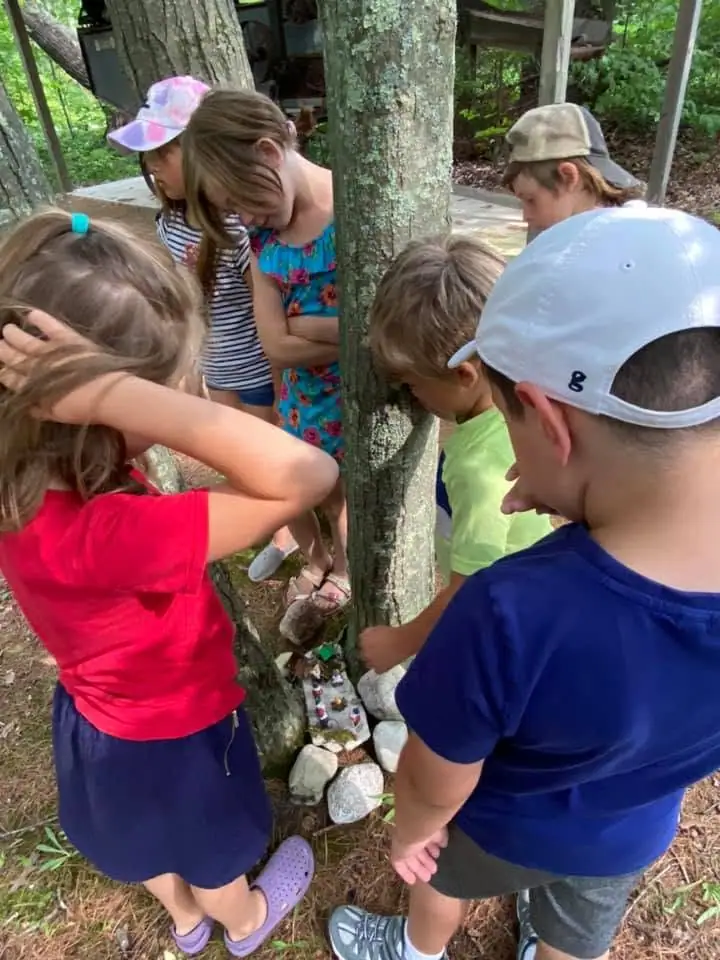 Children gather around a tree to look at a small miniature garden arranged among stones.