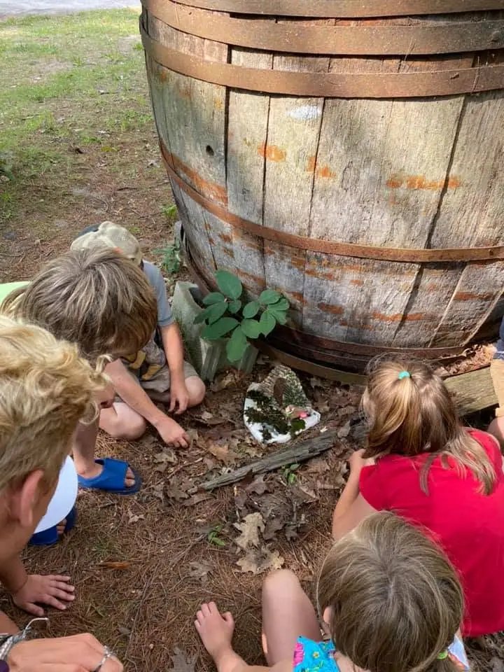 Children gather around a small fairy garden beside a large weathered wooden barrel.