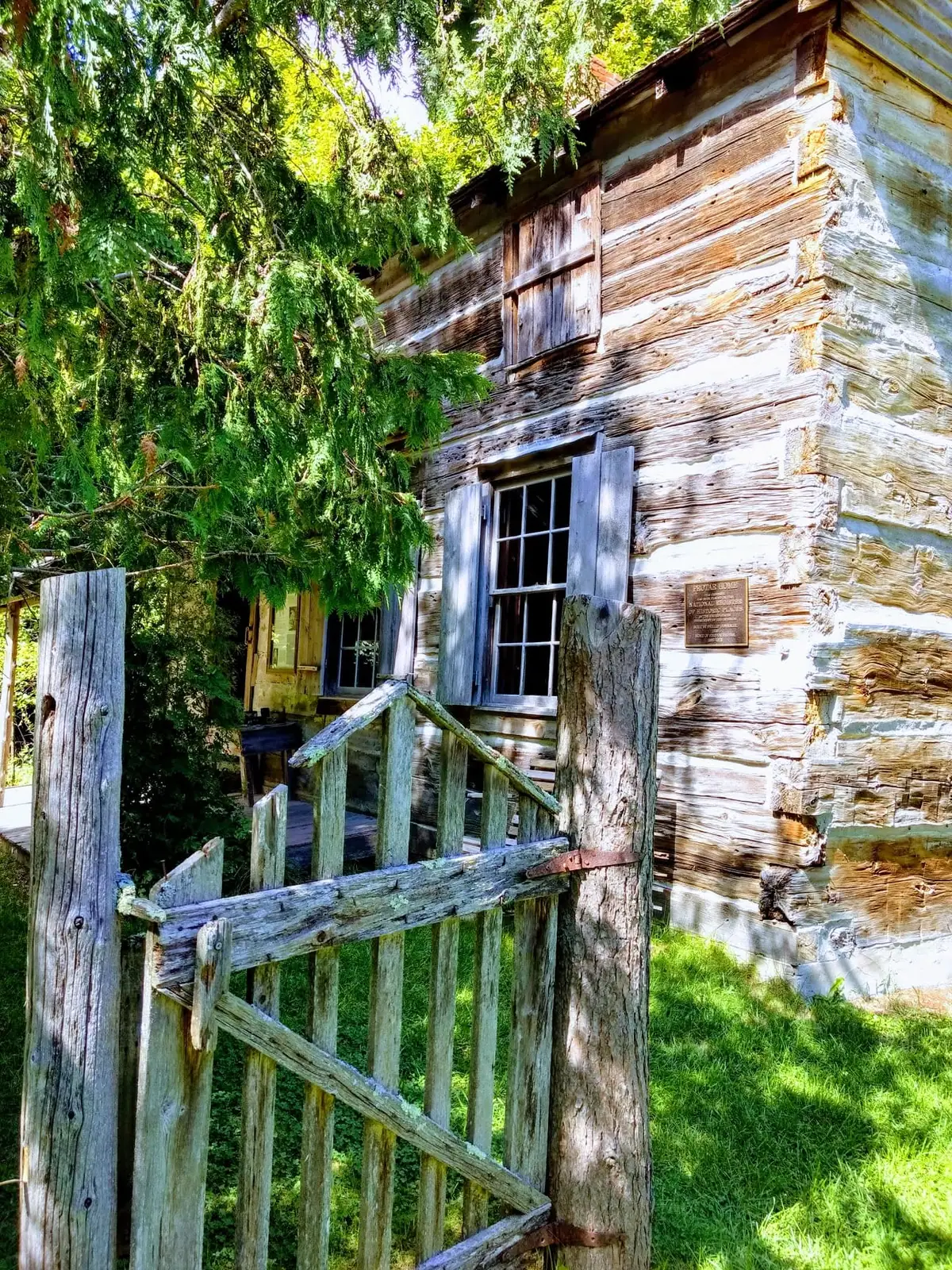 Weathered log house with a small plaque, wooden gate, and fence beneath leafy trees.