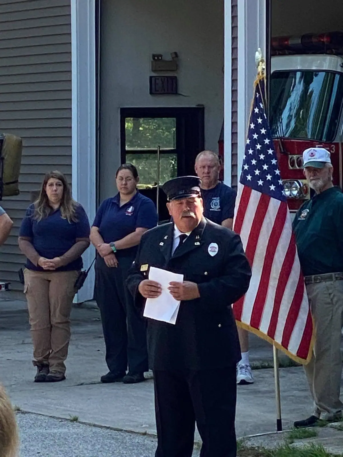 A uniformed man holds a paper before a group beside an American flag and fire truck.