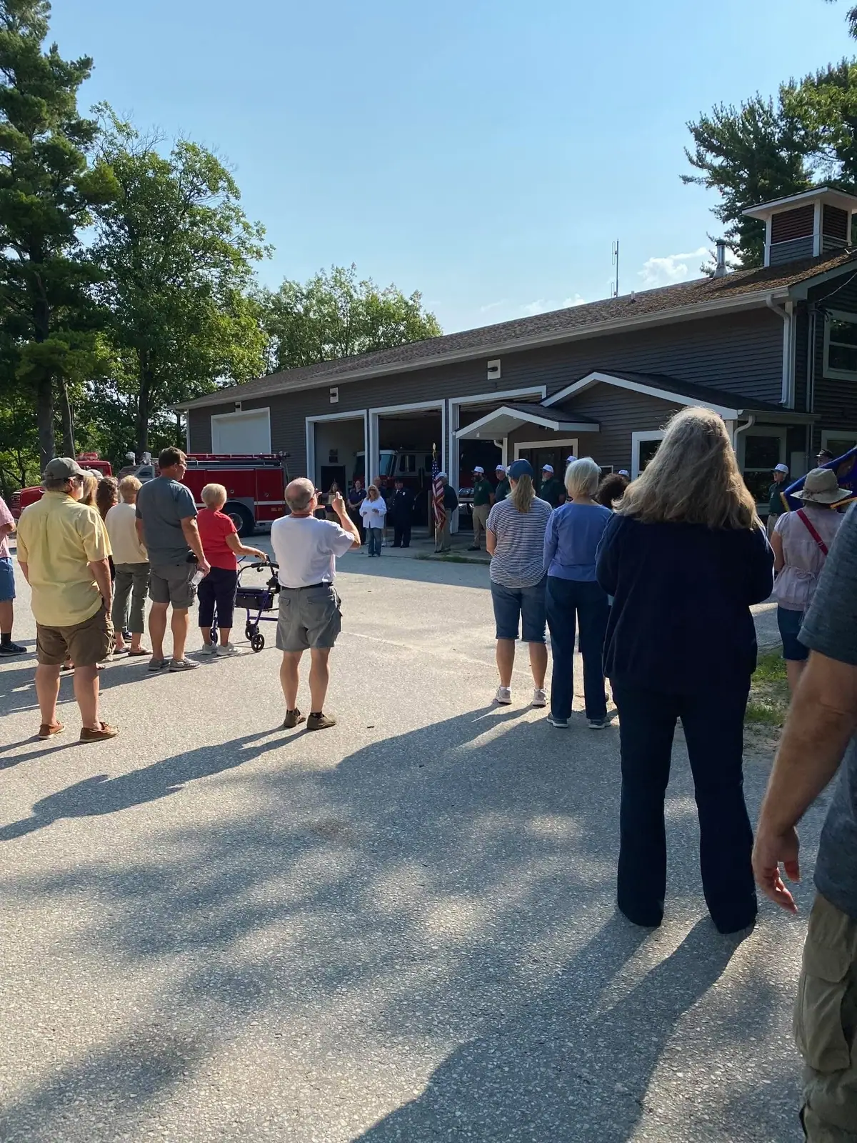A crowd gathers outside a fire station while uniformed personnel stand near an open garage and fire truck.