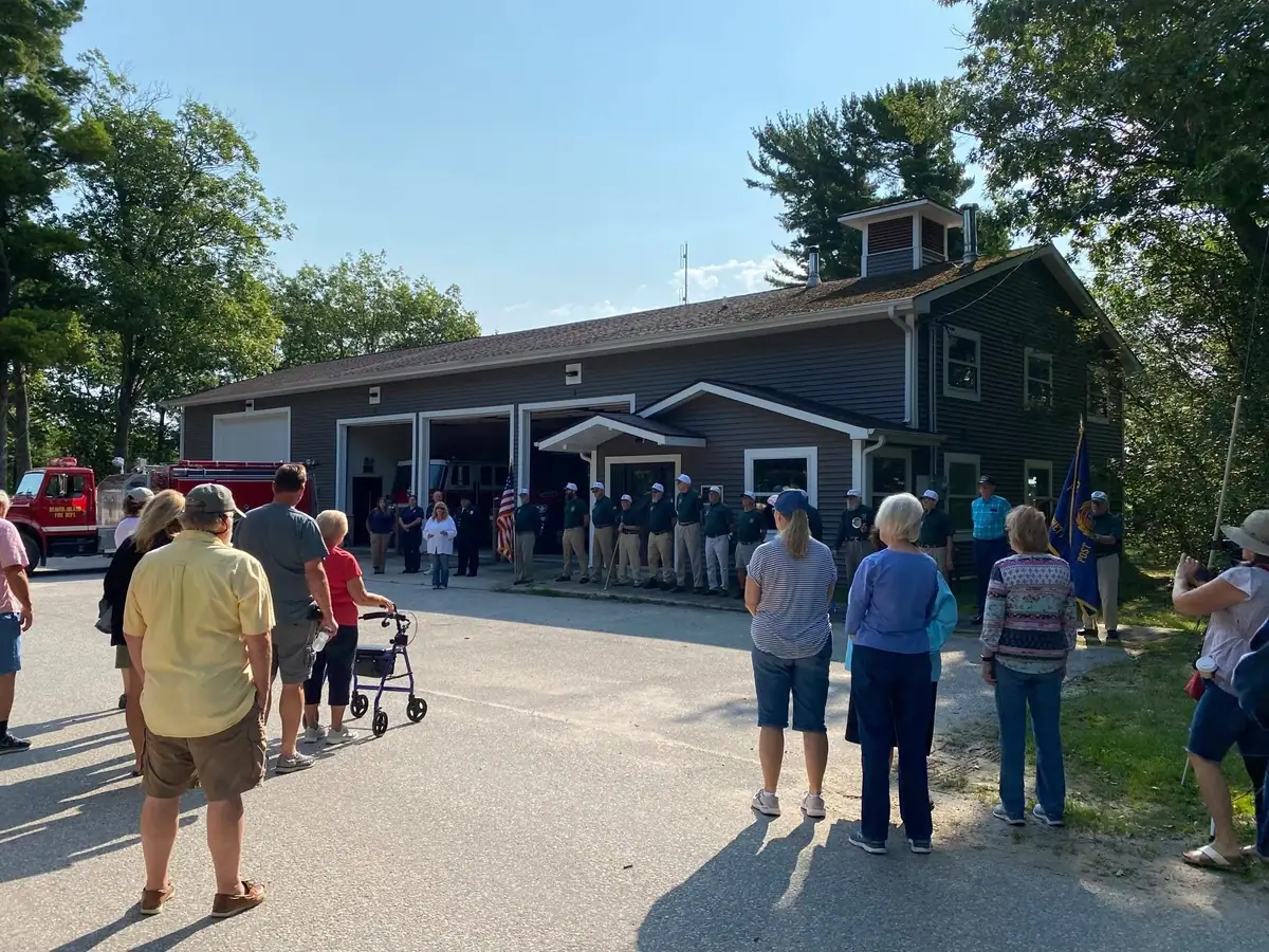A crowd watches uniformed personnel gathered outside a fire station beside a red fire truck.
