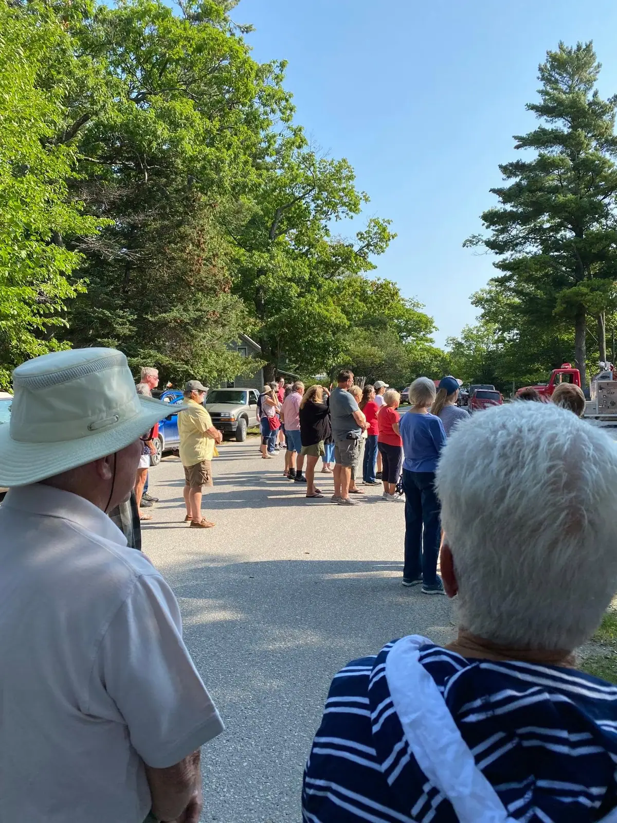 People stand in a line along a tree-lined paved road while others watch from the foreground.