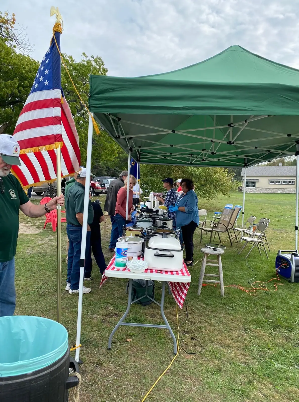 People gather around a food-serving table beneath a green canopy beside an American flag and portable generator.