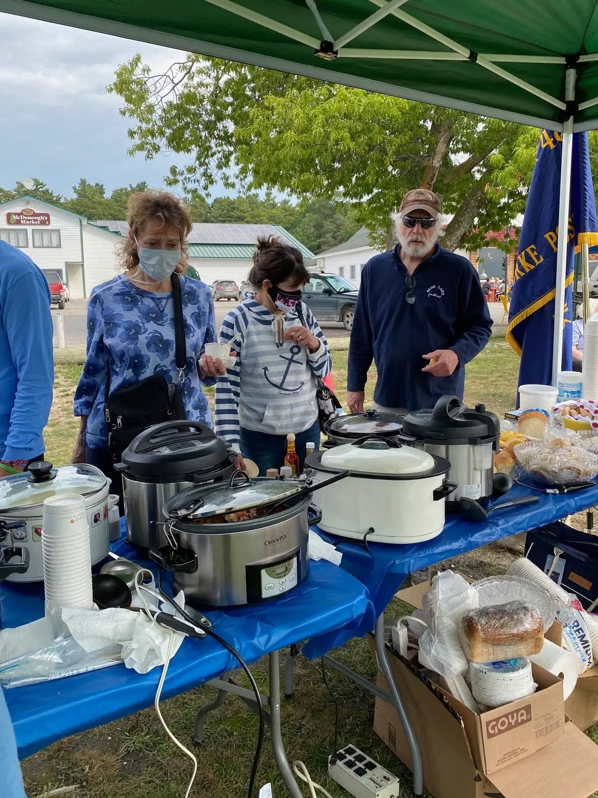 People gather around a food table beneath a green canopy, with slow cookers, cups, bread, and a blue flag visible.