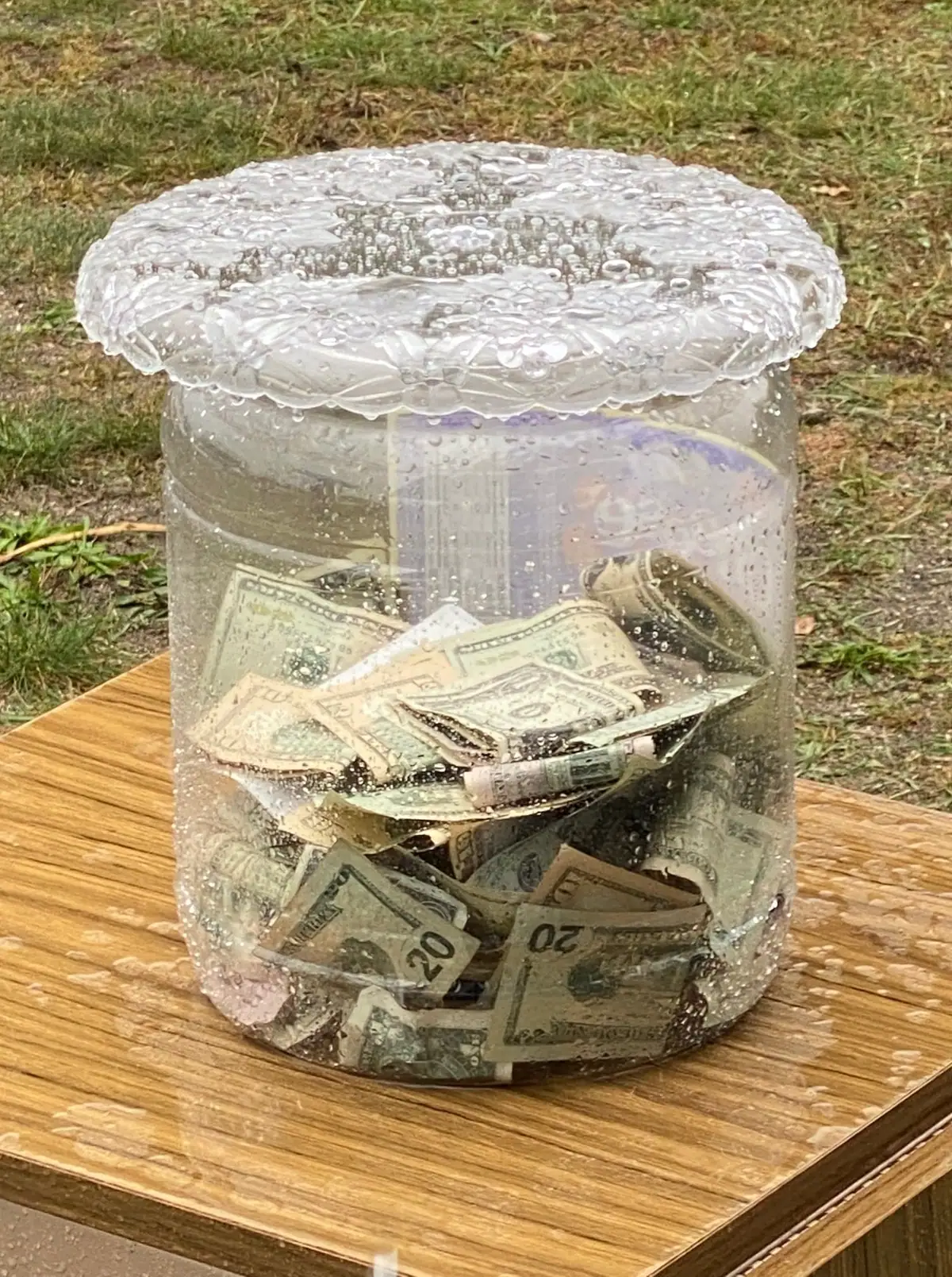 A clear lidded container filled with dollar bills sits on a wet wooden table outdoors.