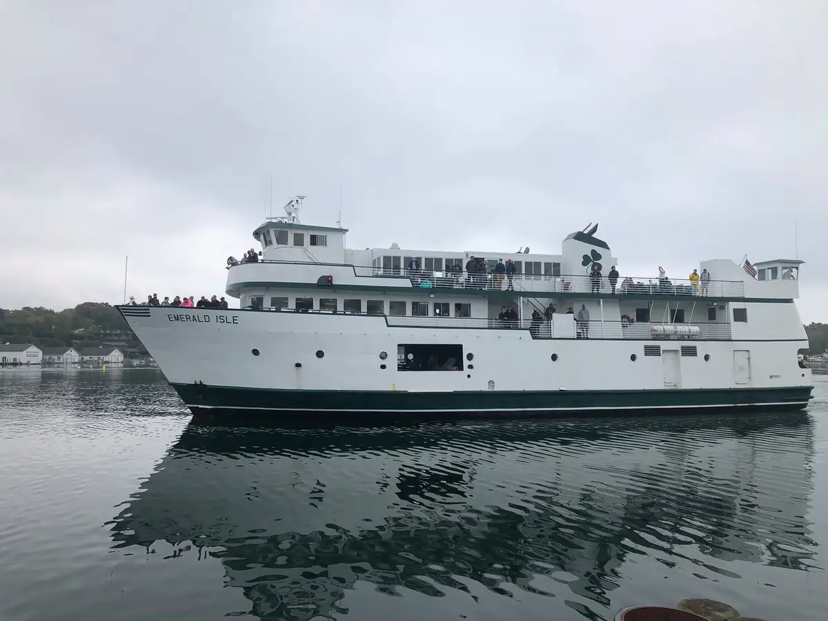 A white passenger ferry carrying people cruises past a wooded shoreline under an overcast sky.