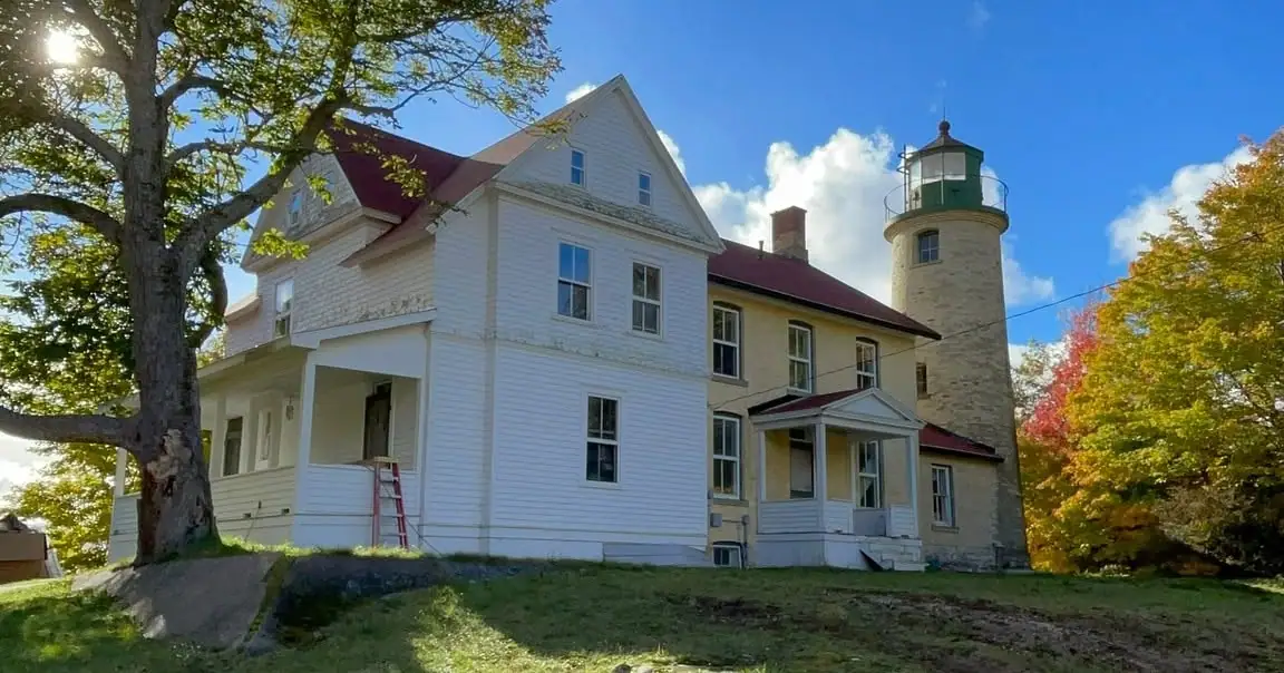 A white lighthouse keeper’s house and round stone light tower stand among autumn trees beneath a blue sky.