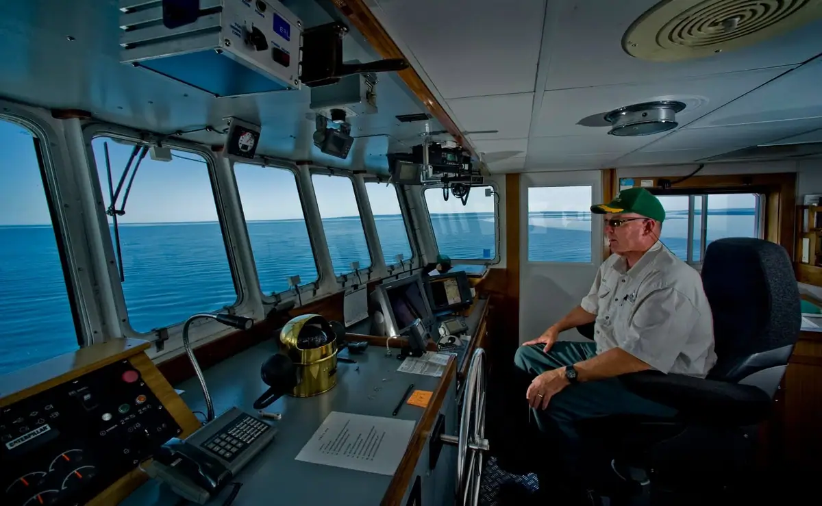 A man sits in a ship’s navigation bridge surrounded by controls and windows overlooking open water.