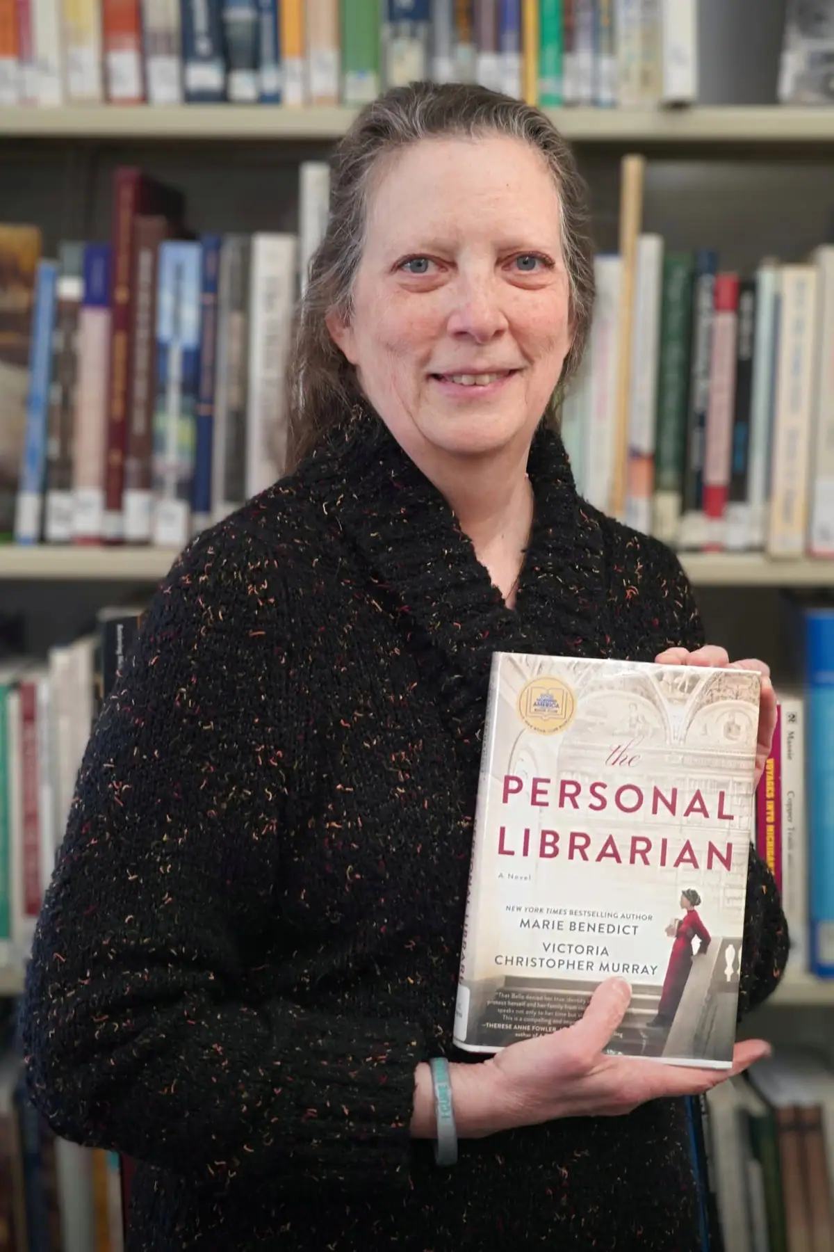 A woman holds a book in front of shelves filled with books.