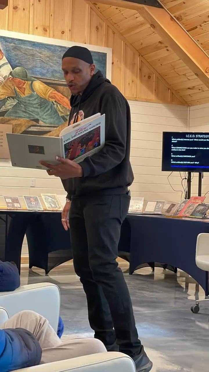A man reads a book while holding a laptop in a wood-paneled room with a mural and display tables.