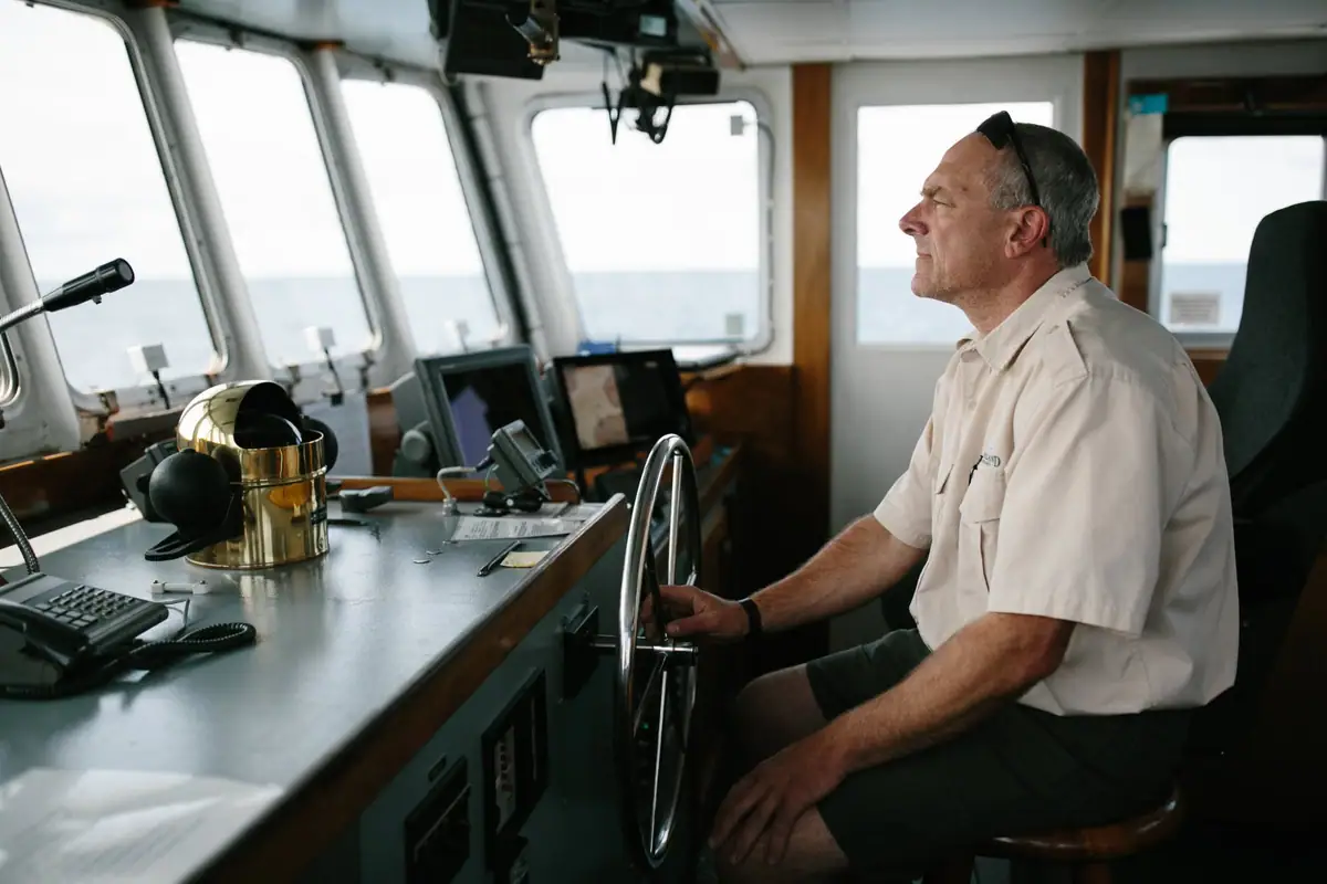 A man sits at a vessel’s helm inside a wheelhouse, surrounded by navigation equipment and looking through the forward windows.