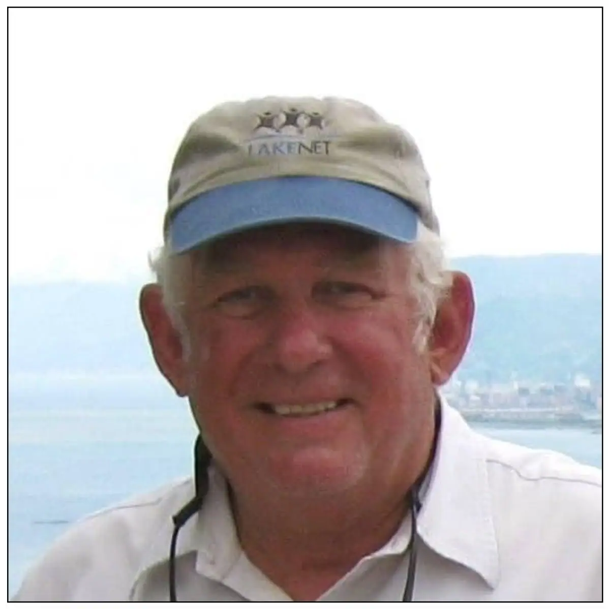 Smiling man in a tan and blue cap and white shirt, photographed outdoors with water and distant hills behind him.