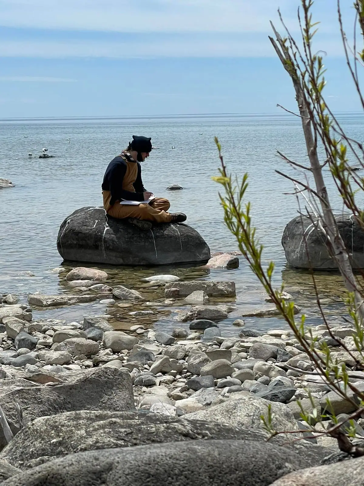 Person writes in a notebook while seated on a rock beside calm water.