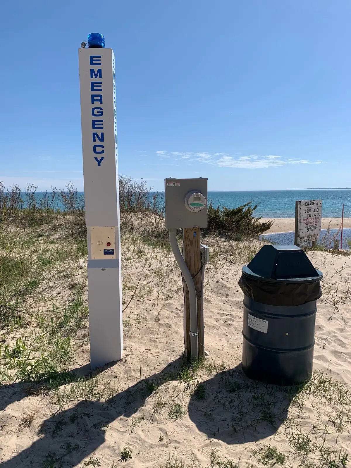 An emergency call tower, utility box, and trash bin stand in sandy dunes beside a beach.