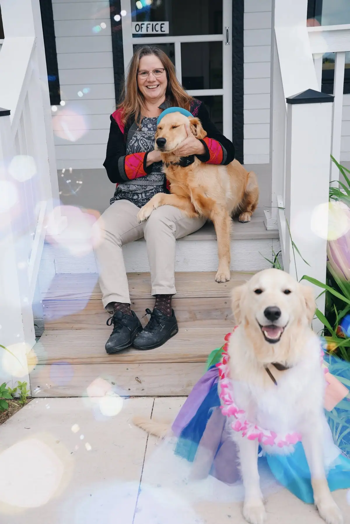 A smiling woman sits on porch steps with two golden retrievers, one wearing a colorful costume.