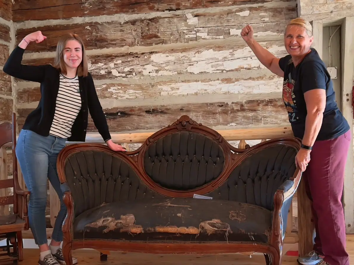 Two women pose with a damaged ornate sofa inside a rustic wood-paneled room.