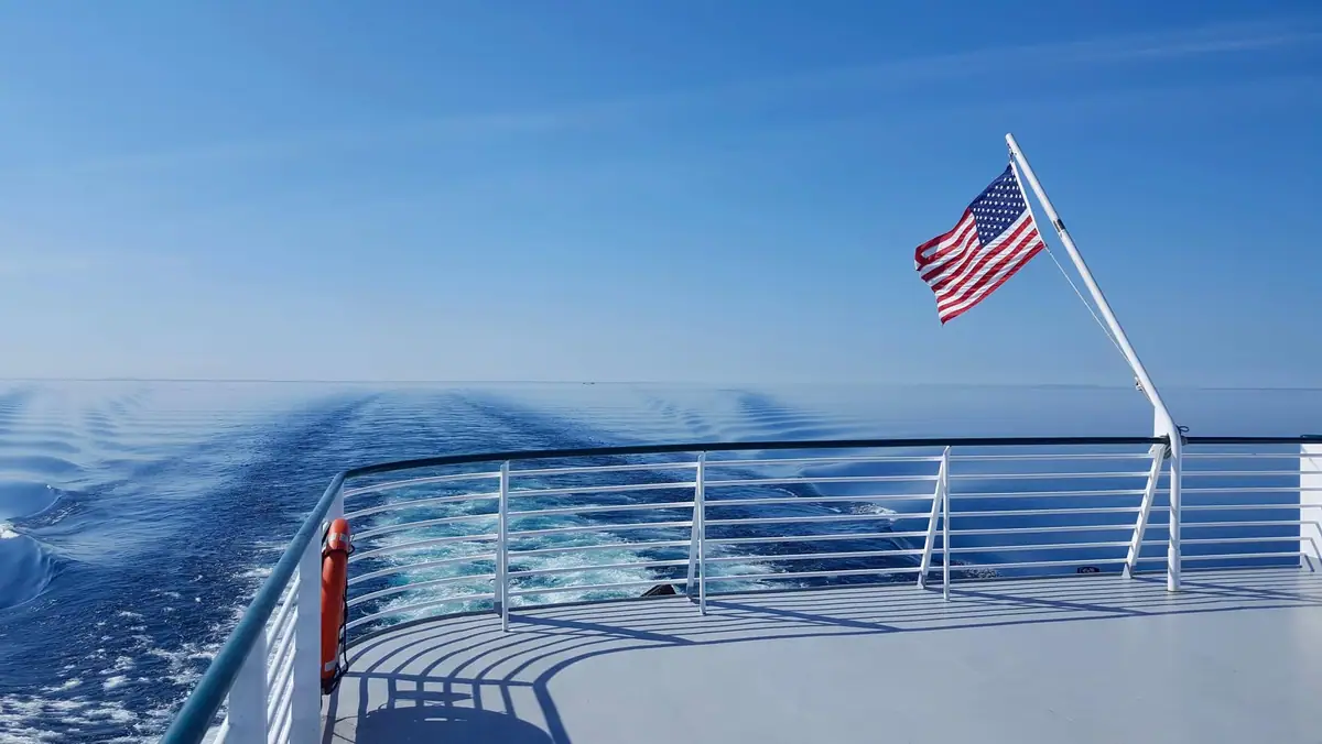 A vessel’s open deck overlooks a foamy wake beneath an American flag.