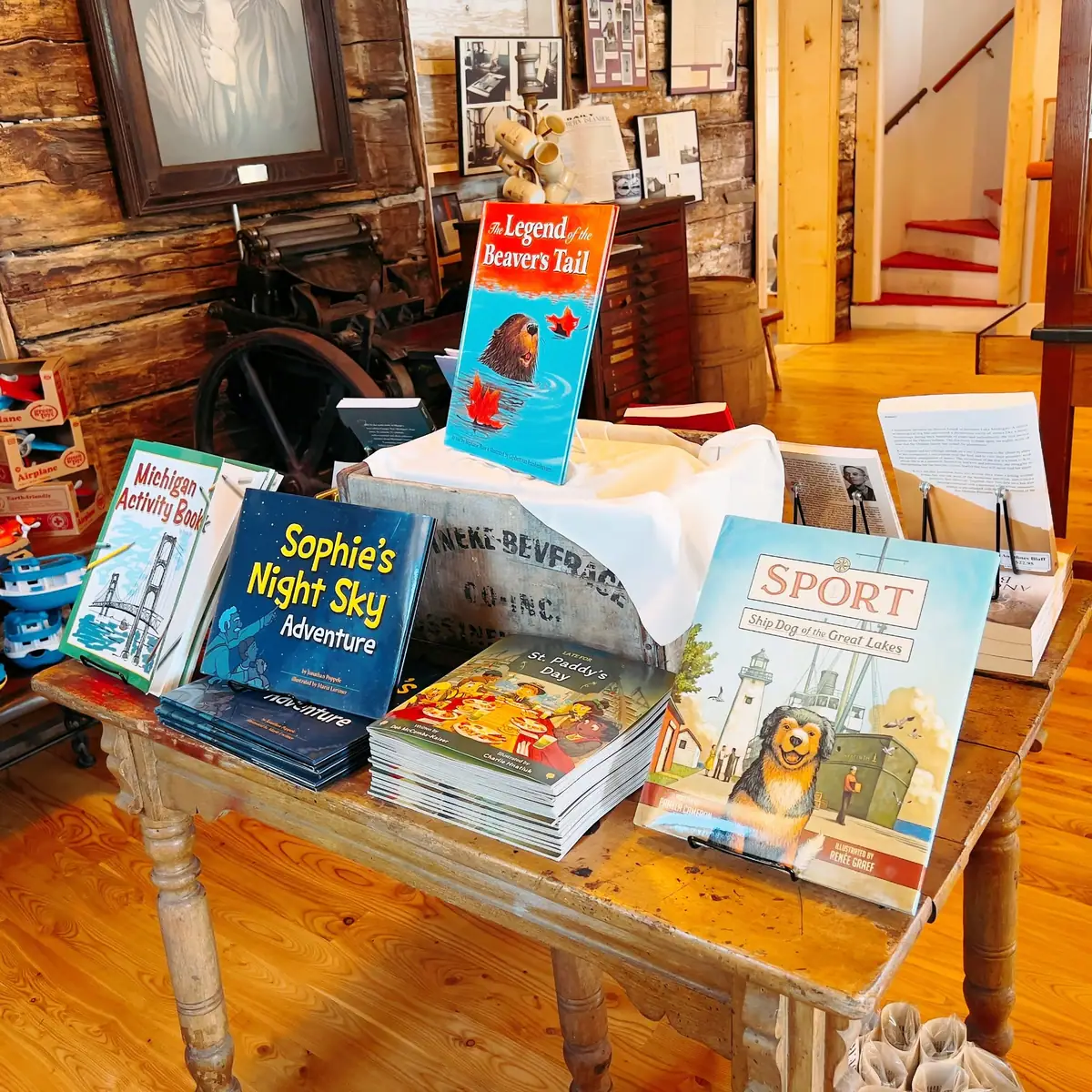 A table displays illustrated books inside a log-walled interior with historic objects and framed materials.