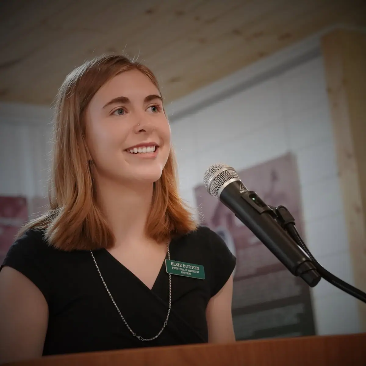 Portrait of a smiling young woman wearing a name badge beside a microphone indoors.