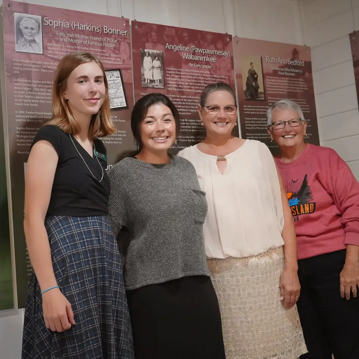 Four women pose in front of historical exhibit panels featuring biographical displays and photographs.