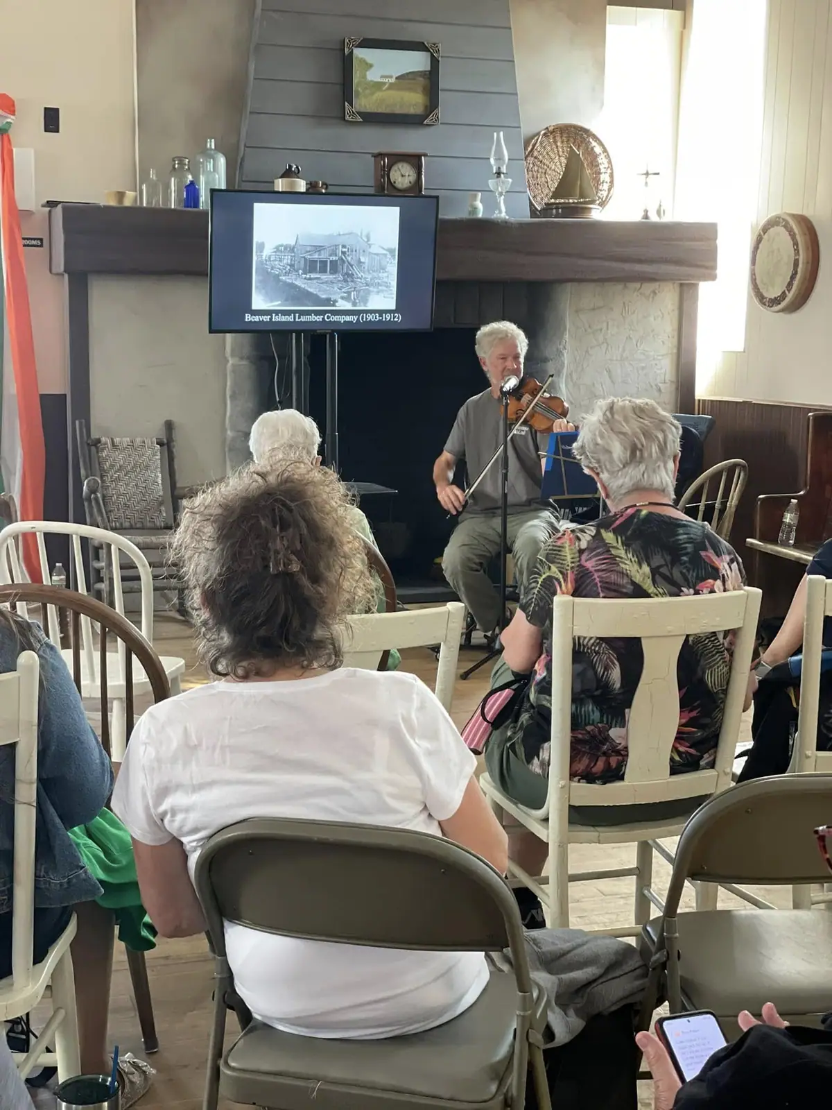 A violinist performs before seated attendees in a room with a stone fireplace and presentation screen.