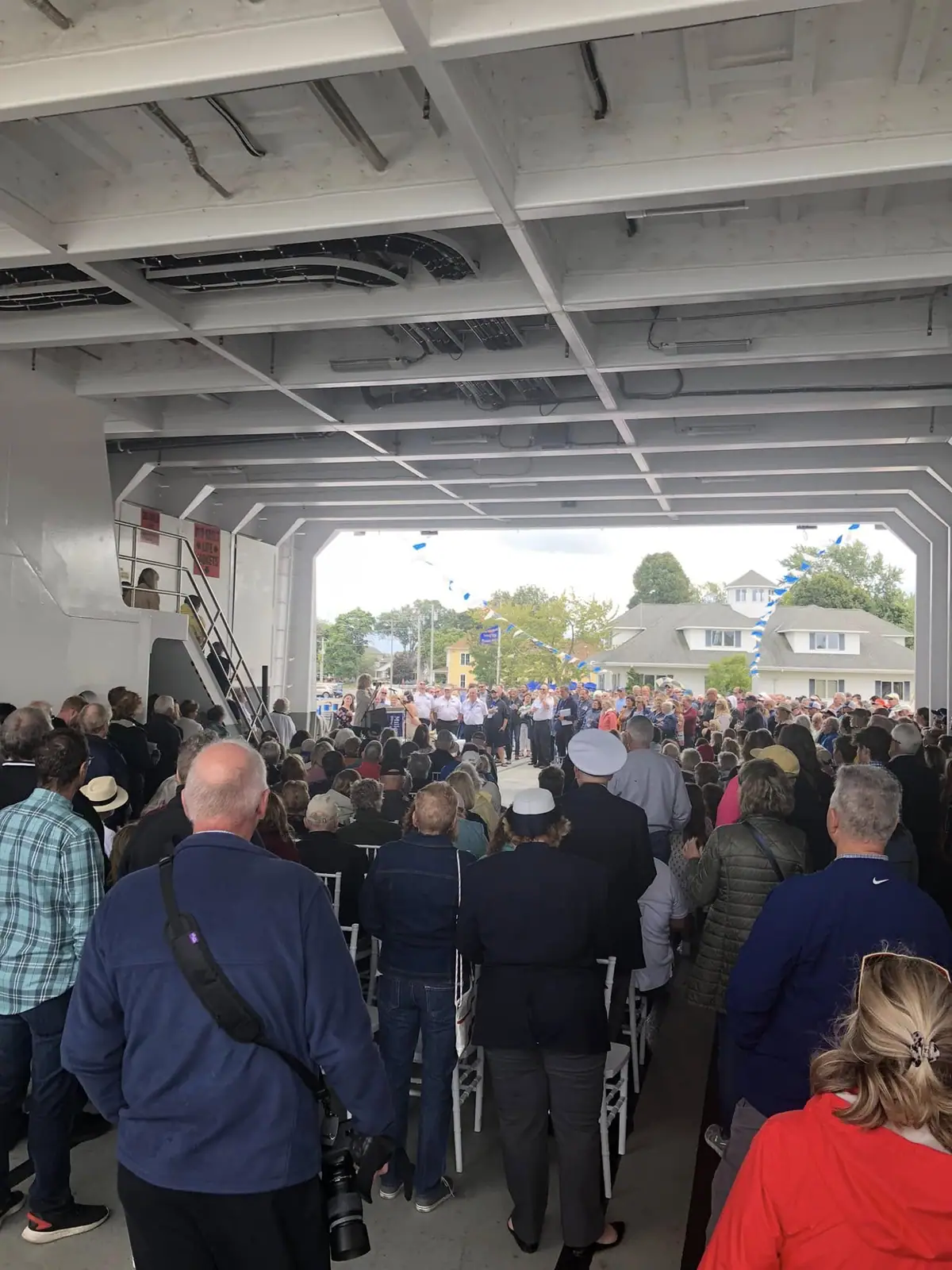 A crowd gathers inside a large vessel beneath blue-and-white bunting while people stand at the open end.