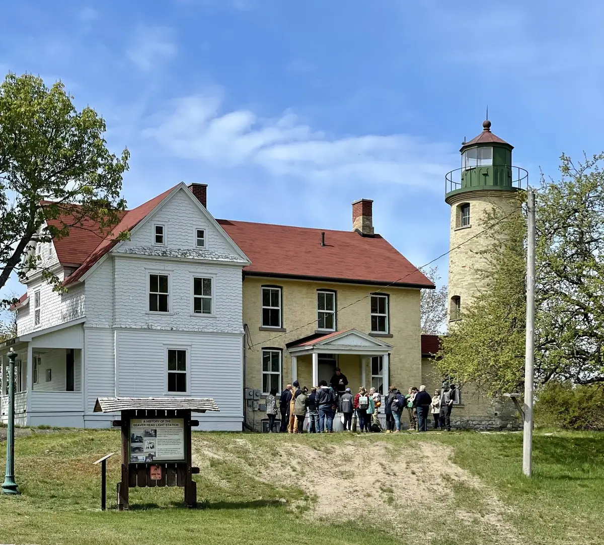 A brick lighthouse tower and adjoining white house stand behind a group gathered near an interpretive sign.
