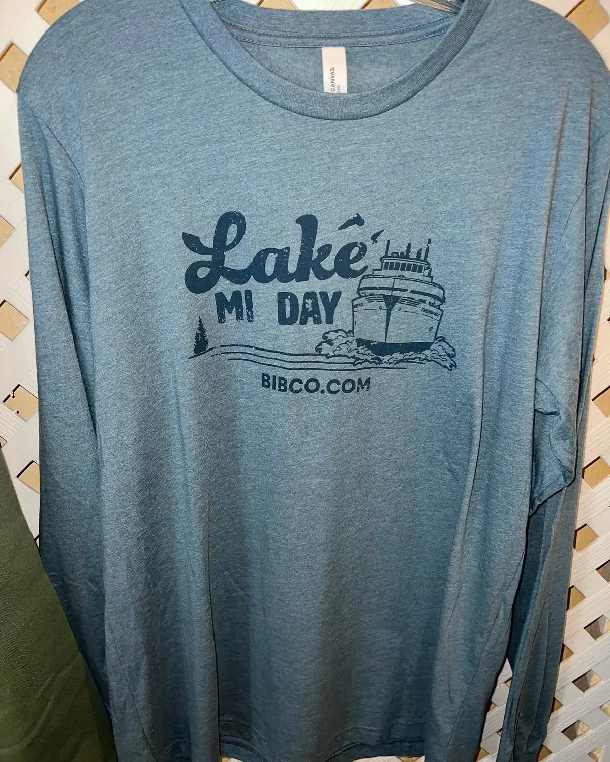 Blue long-sleeved shirt featuring a printed ferry graphic, lettering, an evergreen tree, and flying birds.
