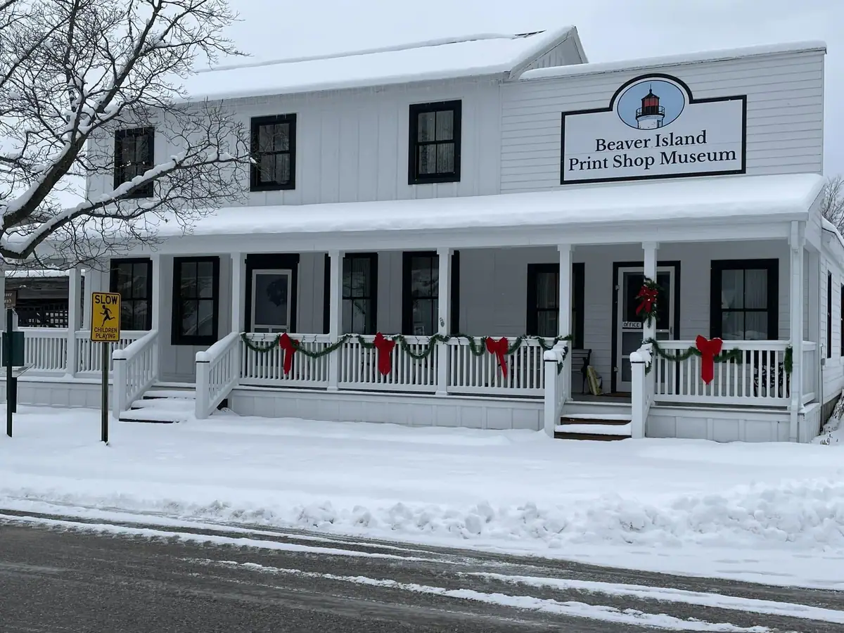 Snow-covered white museum building with a decorated porch, lighthouse sign, and roadside warning sign.