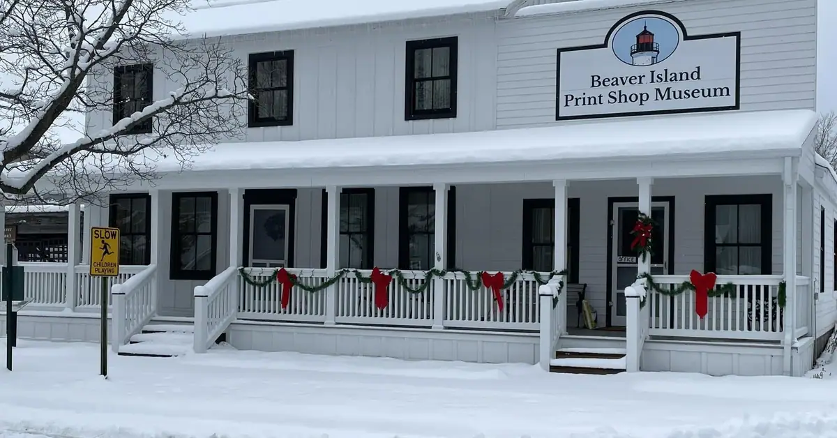 Snow-covered white museum building with holiday garlands, a lighthouse-logo sign, and a roadside warning sign.