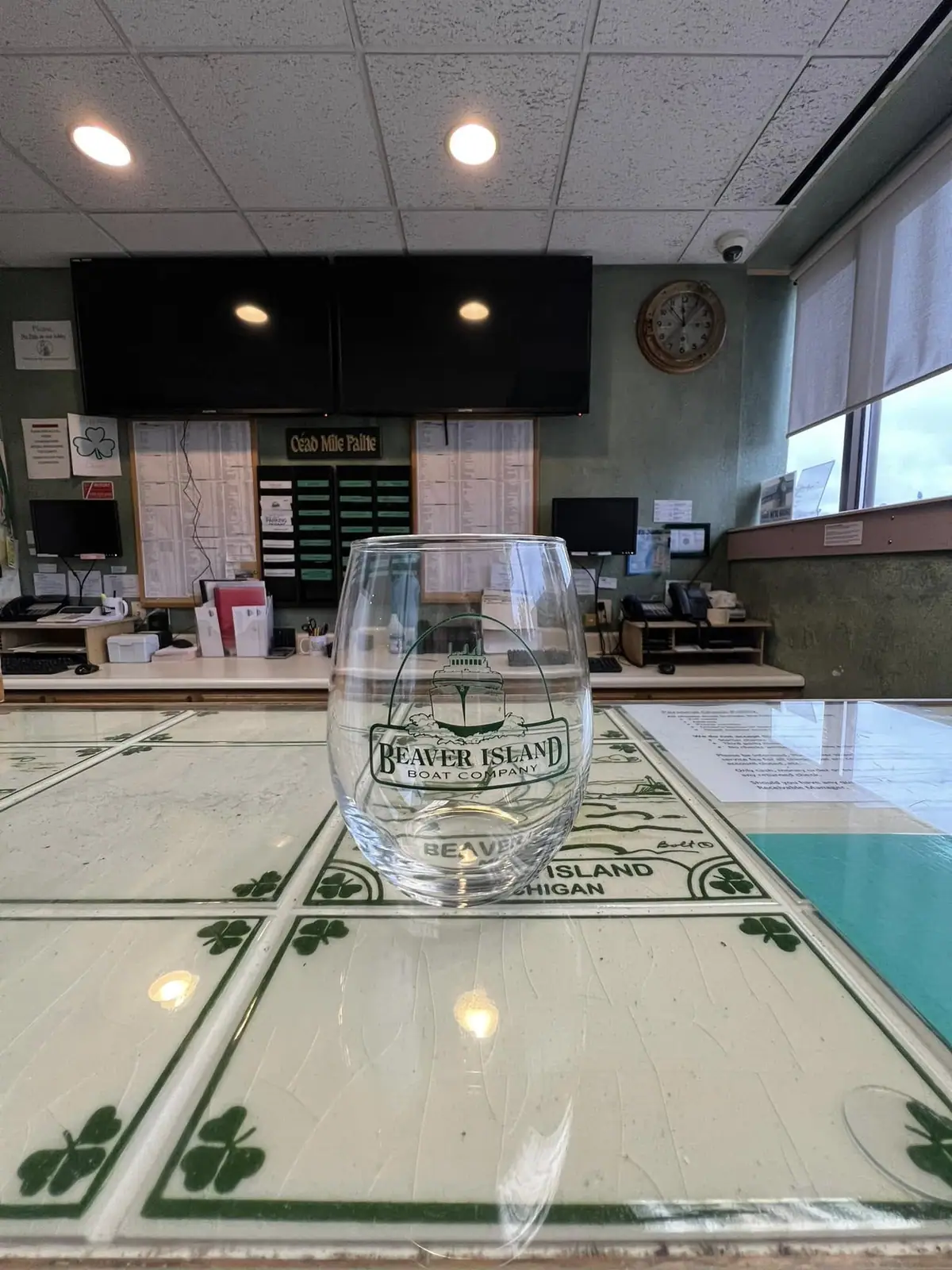 A branded glass sits on a tiled service counter in an office with ticket displays, monitors, and windows.