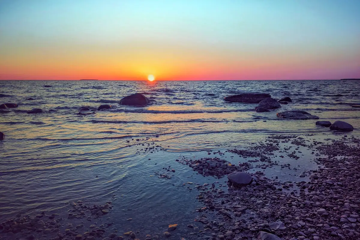 Sunset over shallow water and a rocky shoreline with rounded stones and boulders.