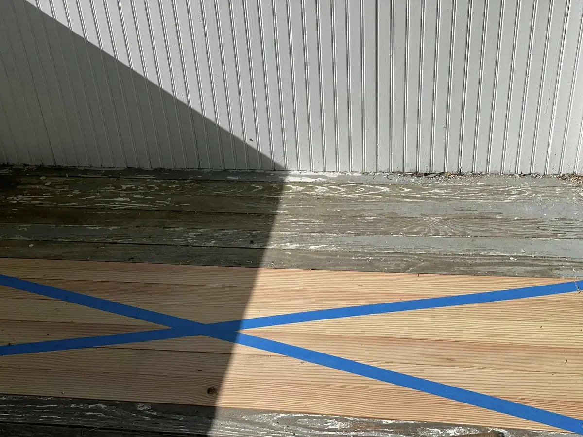 Blue tape forms a large X across wooden floorboards beside a white corrugated wall.