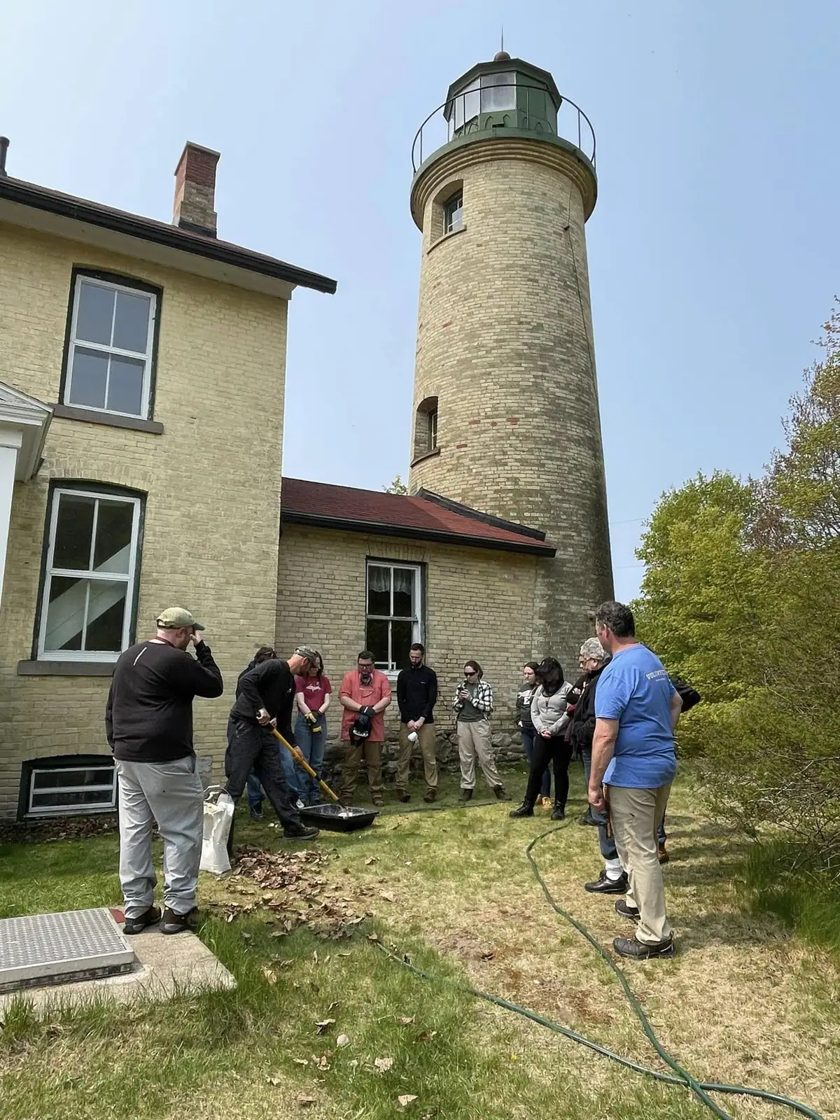 People gather beside a brick lighthouse and adjoining building as one person works on the grounds.