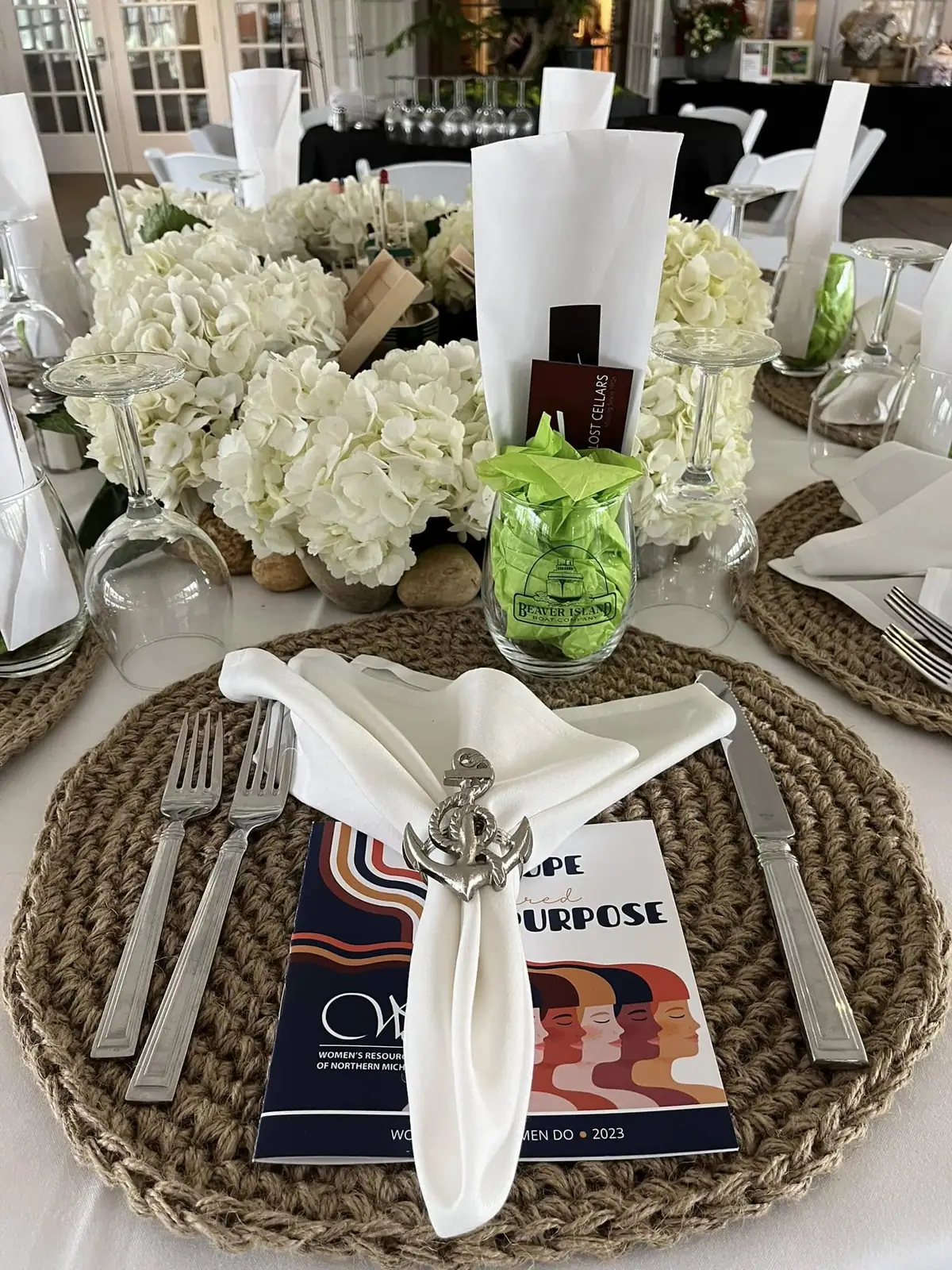 Formal dining table with white floral centerpieces, place settings, wine glasses, and an event booklet.