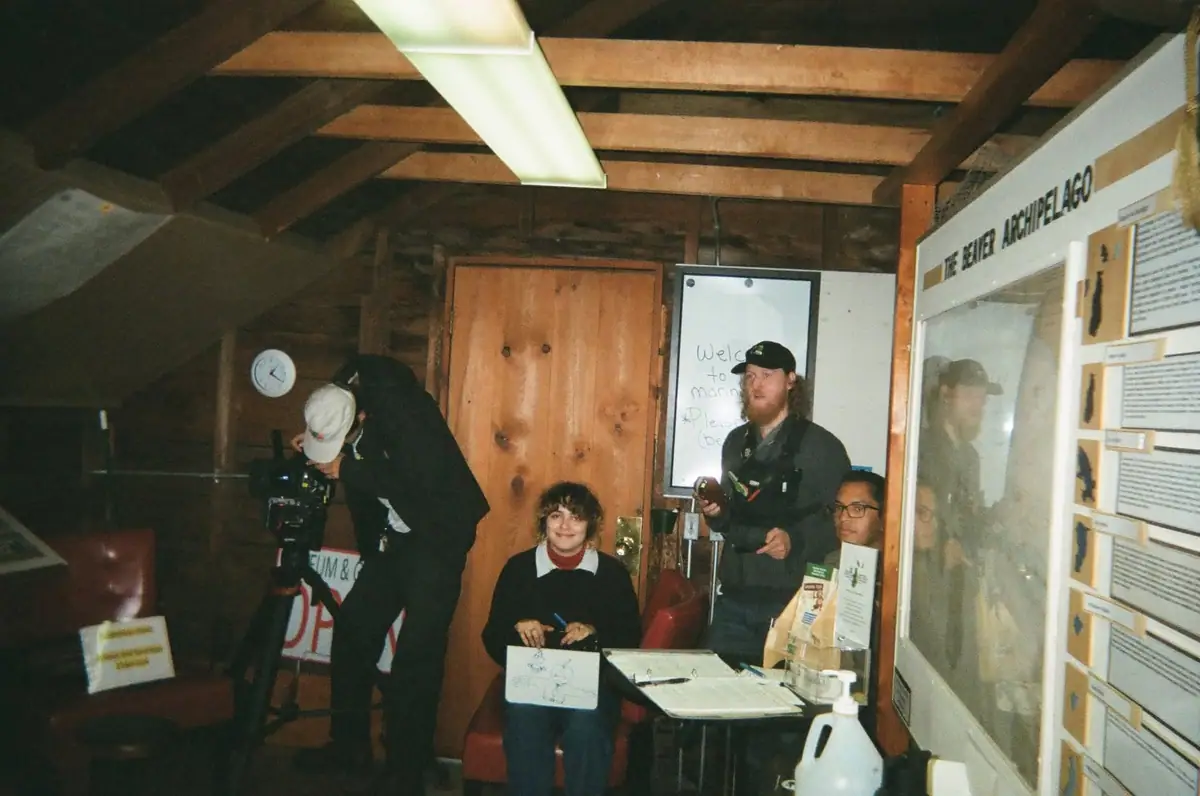 People gather inside a wooden museum room beside exhibit displays and a camera operator.