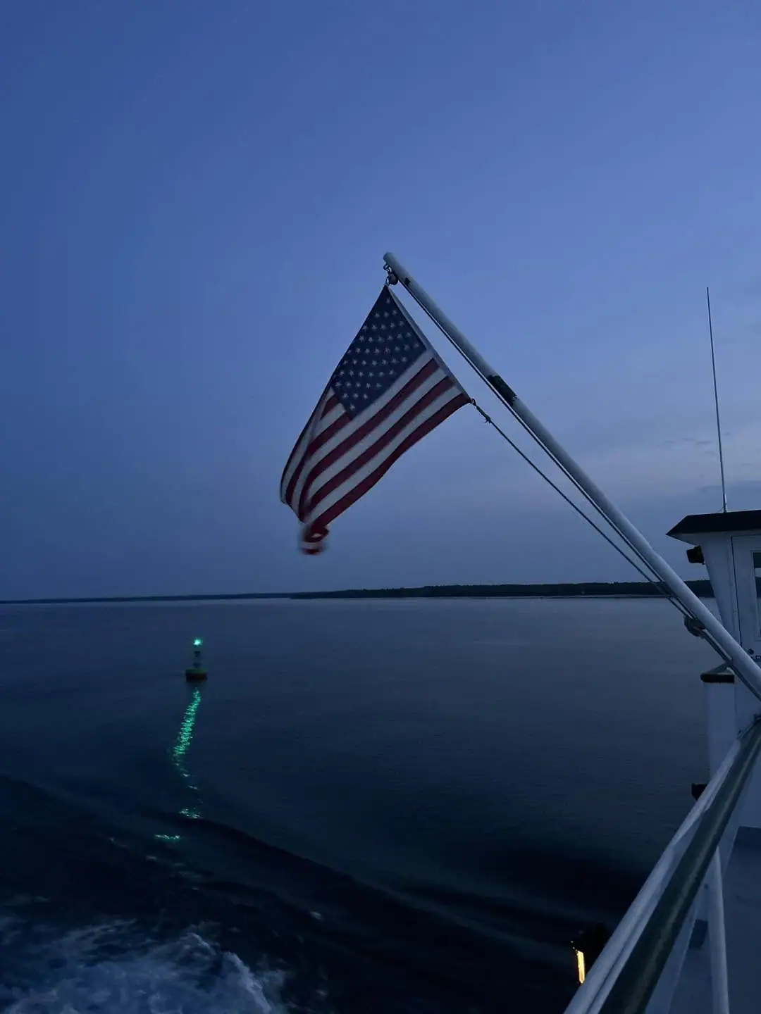 American flag flying from a vessel at dusk beside a green-lit buoy.