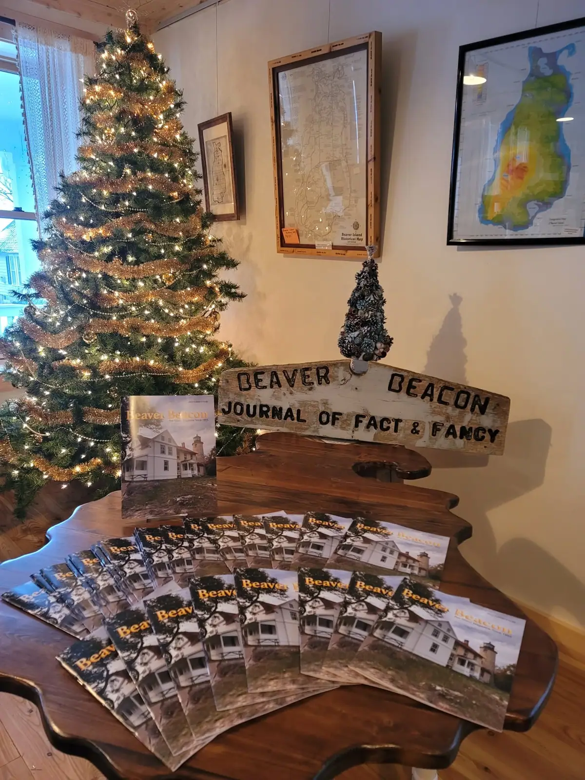 Holiday display with a lit Christmas tree, magazines, a wooden sign, and framed maps in an indoor room.