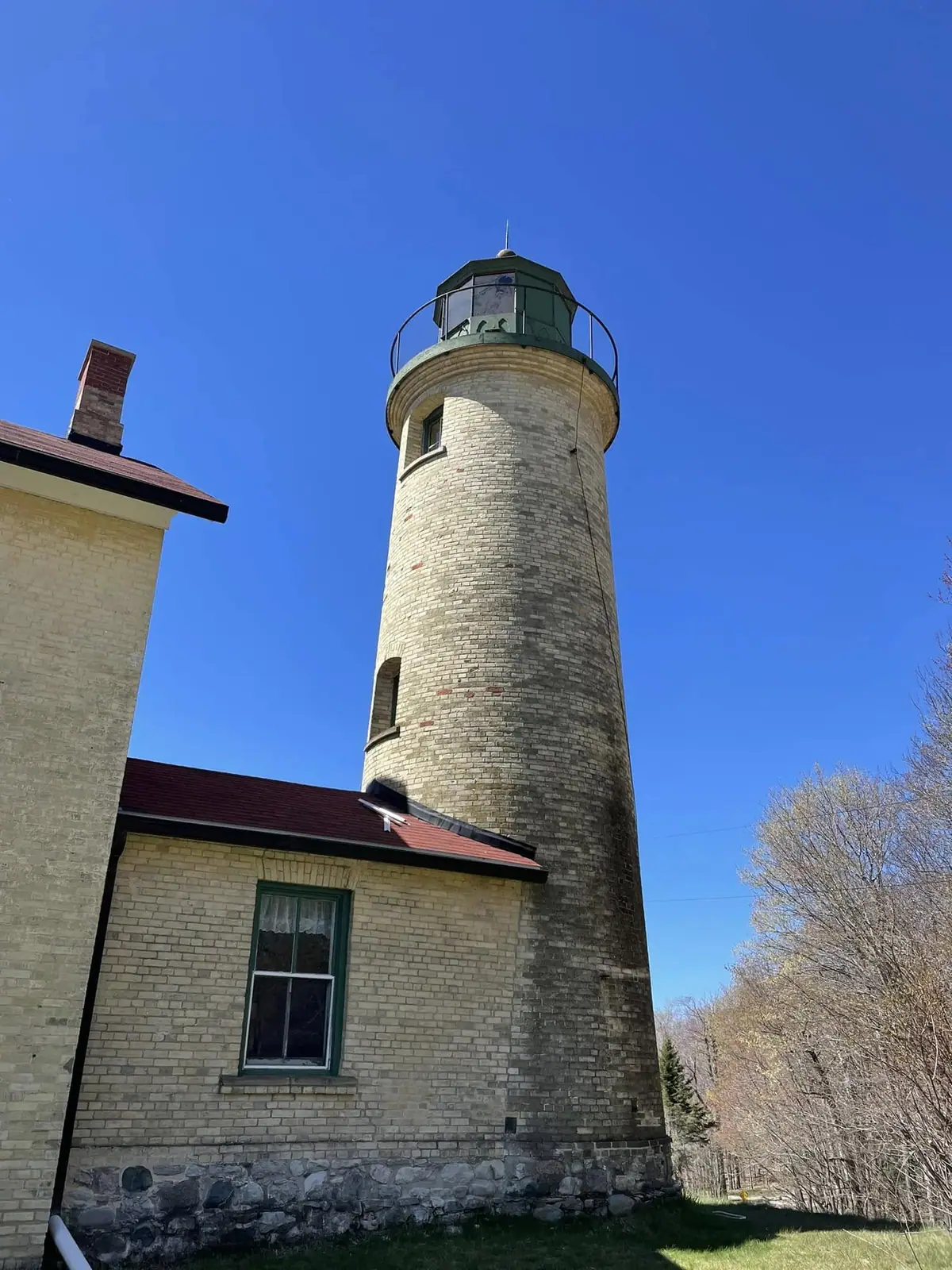 A cylindrical brick lighthouse tower and attached brick building stand beneath a clear blue sky.